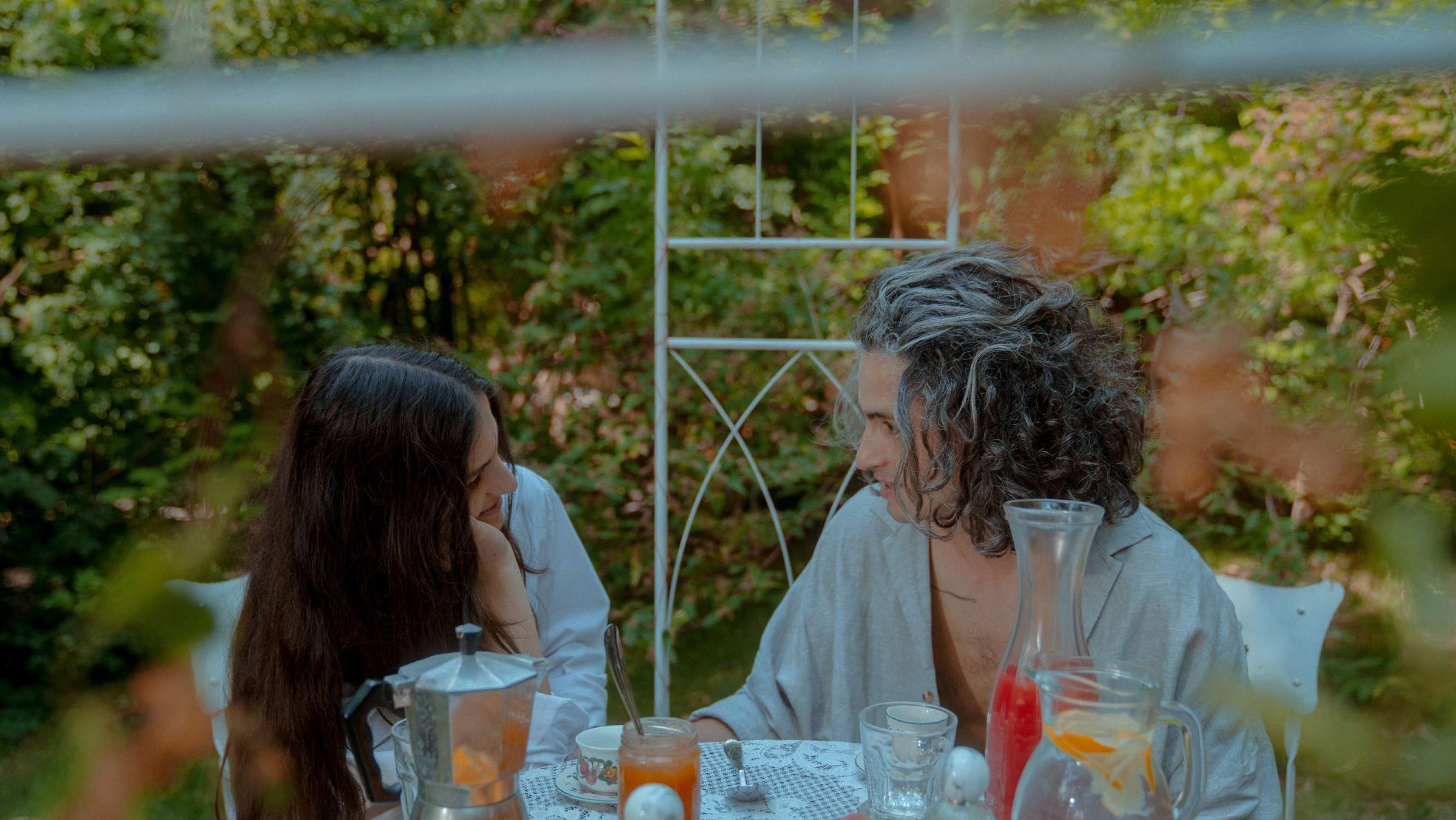 Two artists sit at a table in the garden with drinks, fruit and bottle of Place des Lices perfume.